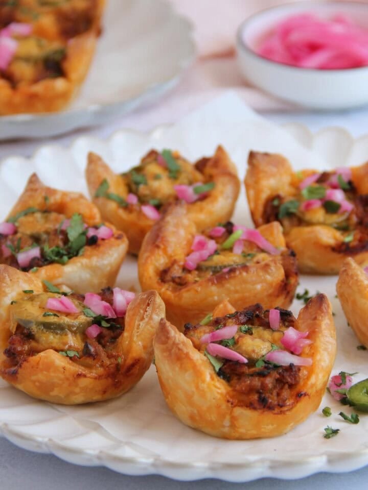 A plate of puff pastry cups filled with a savory mixture and garnished with chopped herbs and pickled onions, with more pastry cups and a bowl of pickled onions in the background.