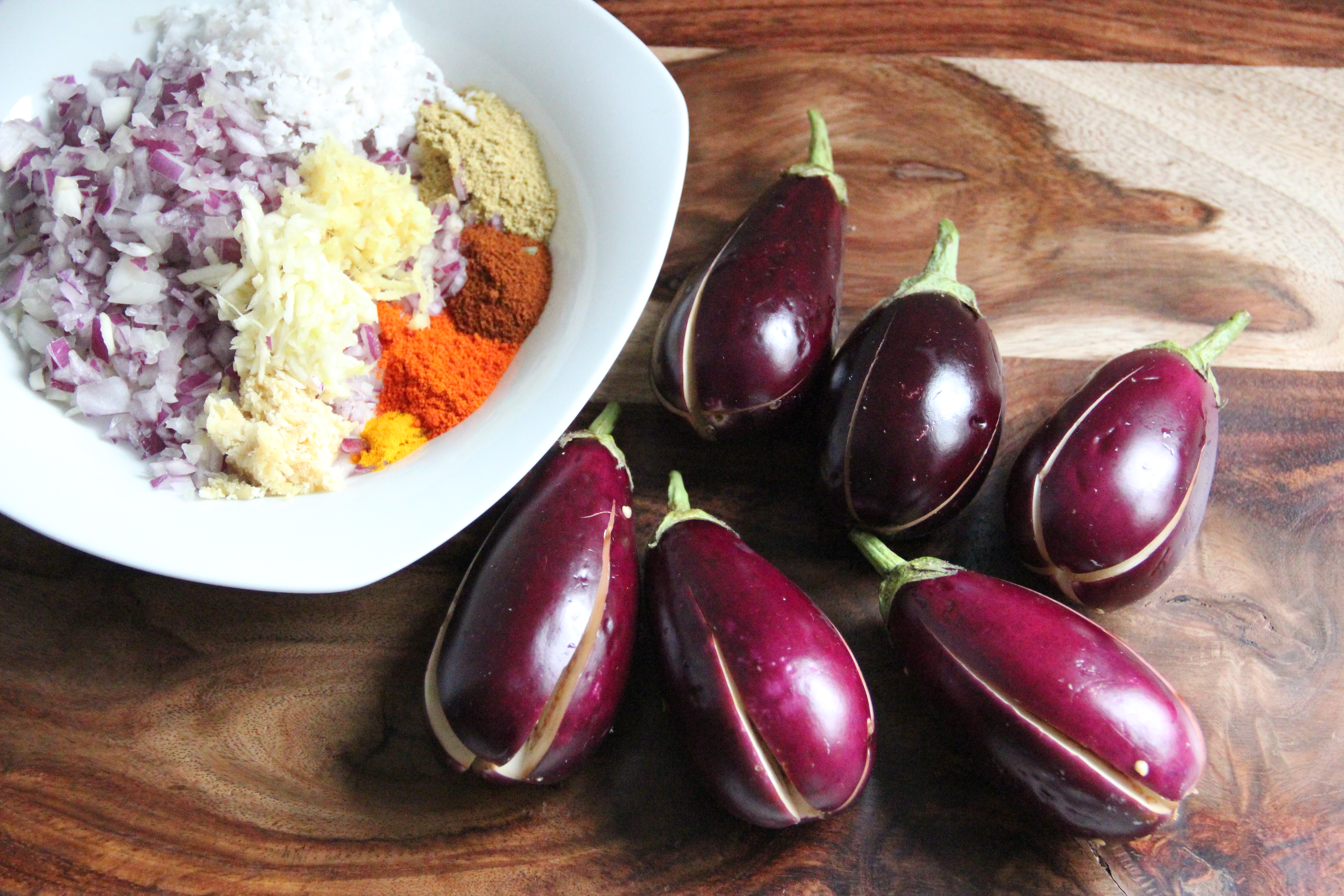 baby eggplants on a wooden cutting board with the ingredients for stuffing in a white plate on teh side