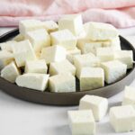 A plate filled with neatly arranged cubes of white paneer cheese, with a few pieces scattered on the white surface next to the plate.