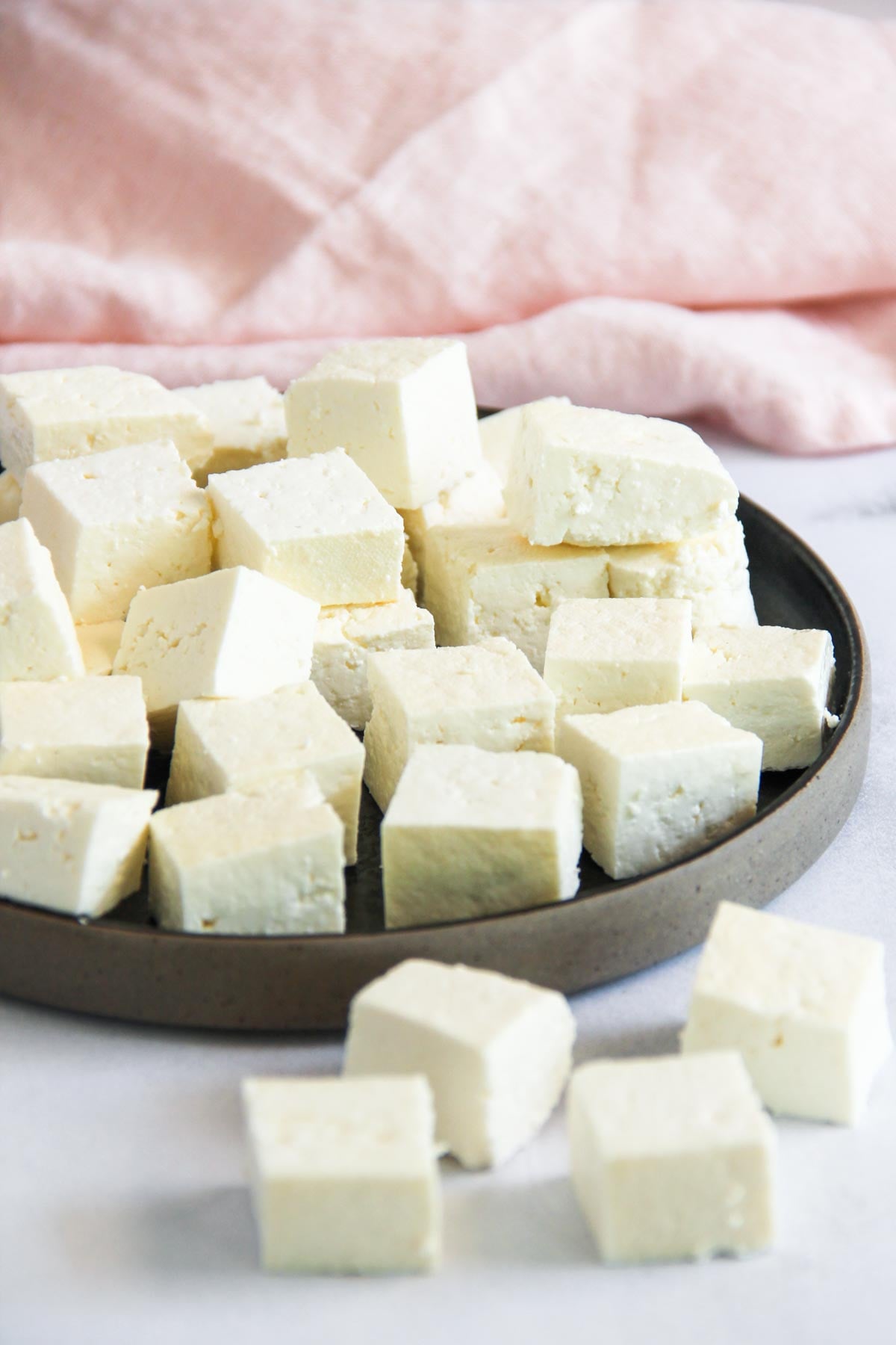 A brown plate holds several cubes of white paneer cheese, with a pink cloth in the background and a few cubes on the surface in front.