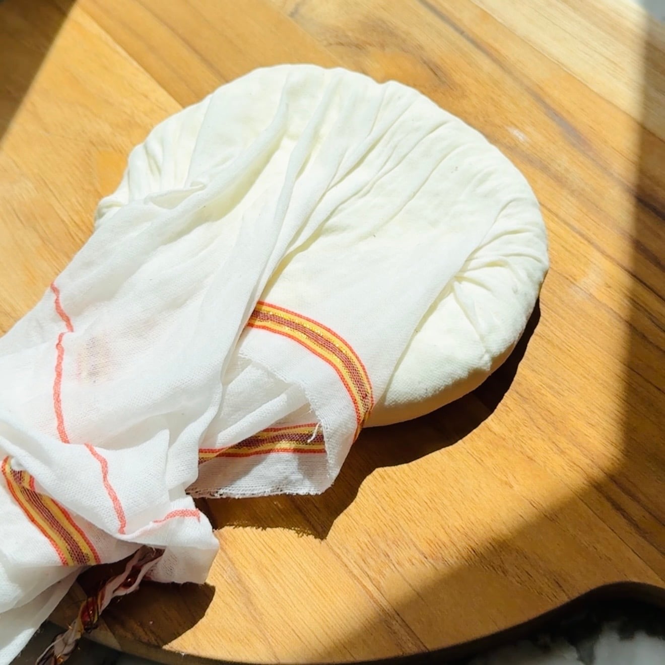 Round ball of white cheese wrapped in a cheese cloth, resting on a wooden surface.