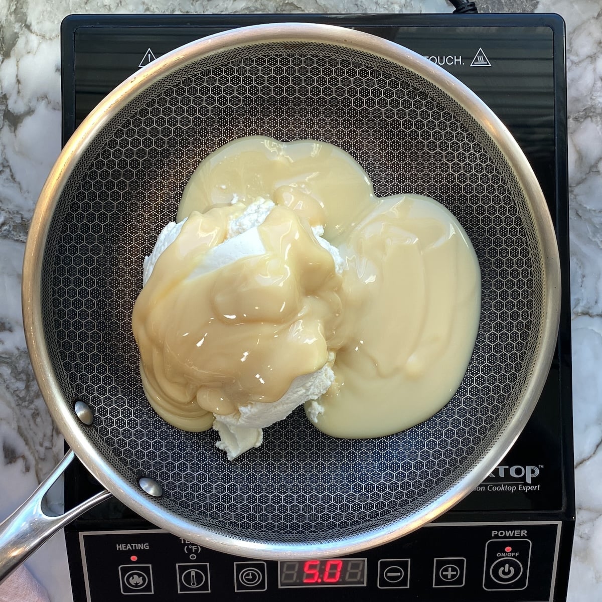 A frying pan on an induction cooktop contains cream cheese and sweetened condensed milk, about to be heated.
