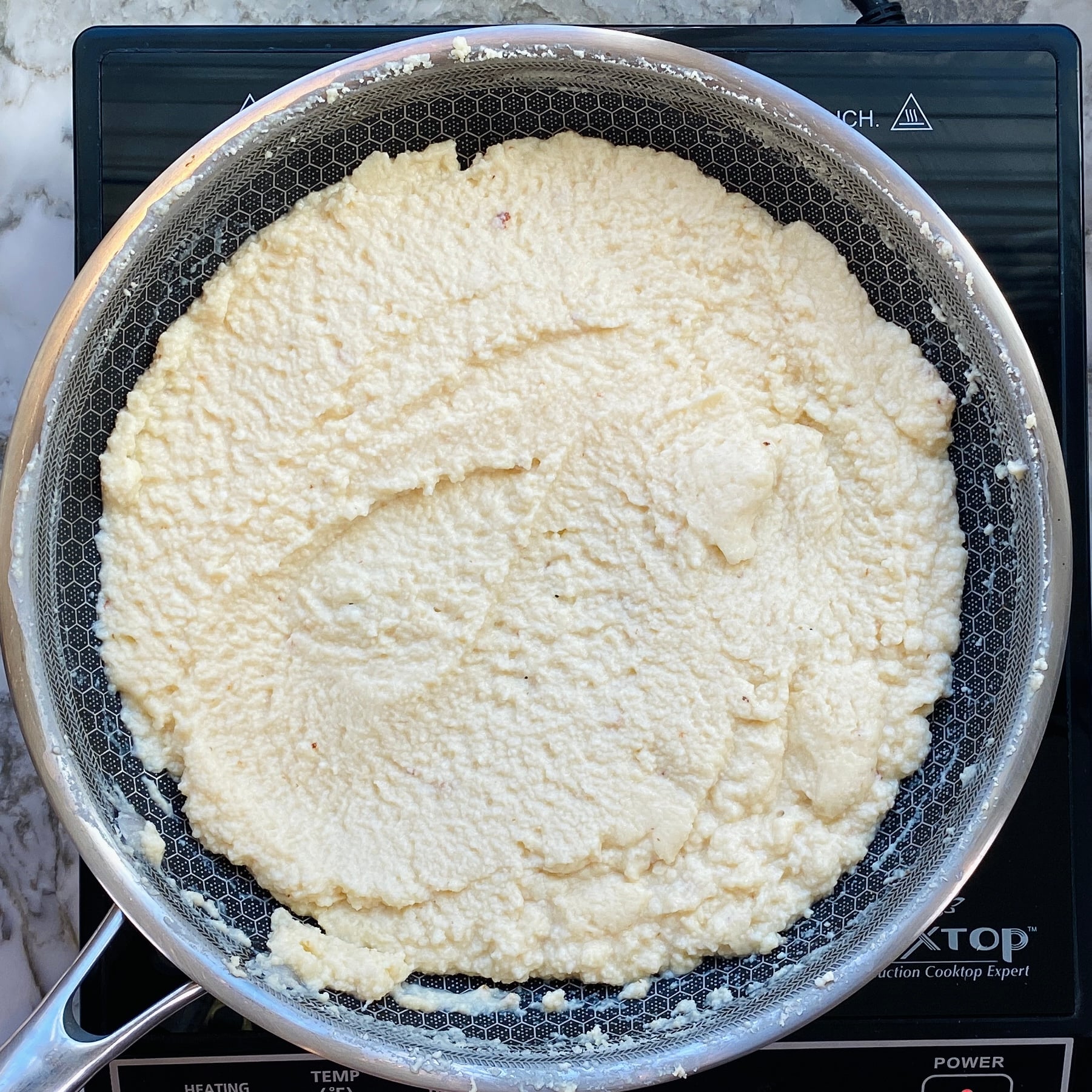 A metal pan on an induction cooktop contains a thick, pale, grainy mixture spread evenly across the surface.