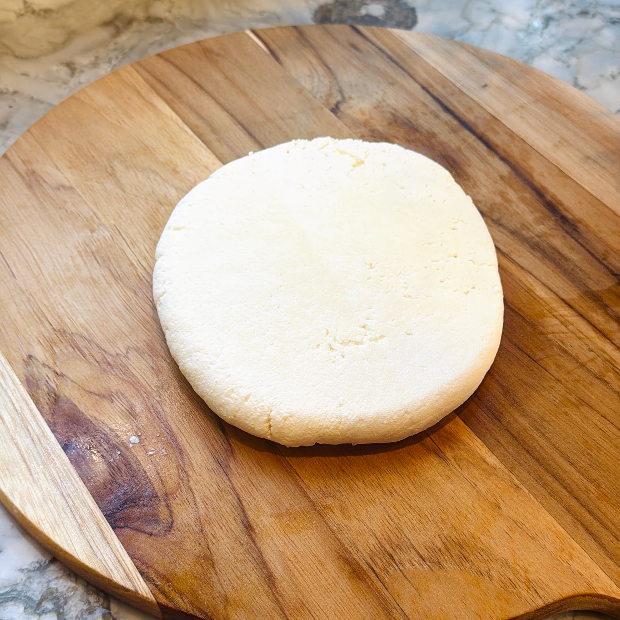 A round, flattened disk of fresh white cheese sits on a wooden cutting board, with a marble countertop in the background.