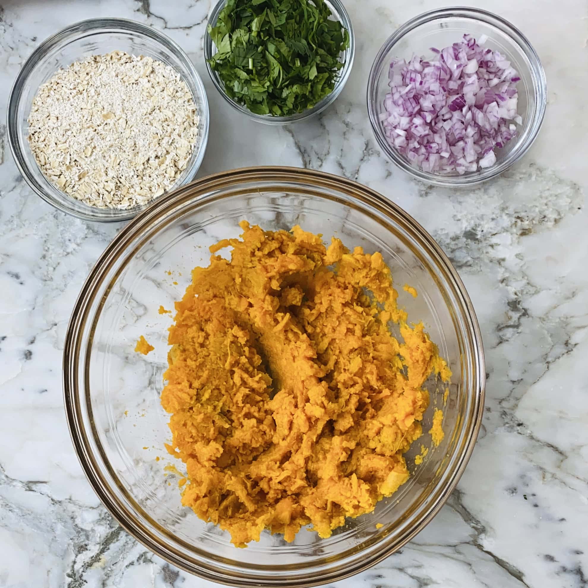 A glass bowl with mashed sweet potato, surrounded by small bowls of chopped red onion, oats, and chopped herbs on a marble countertop.