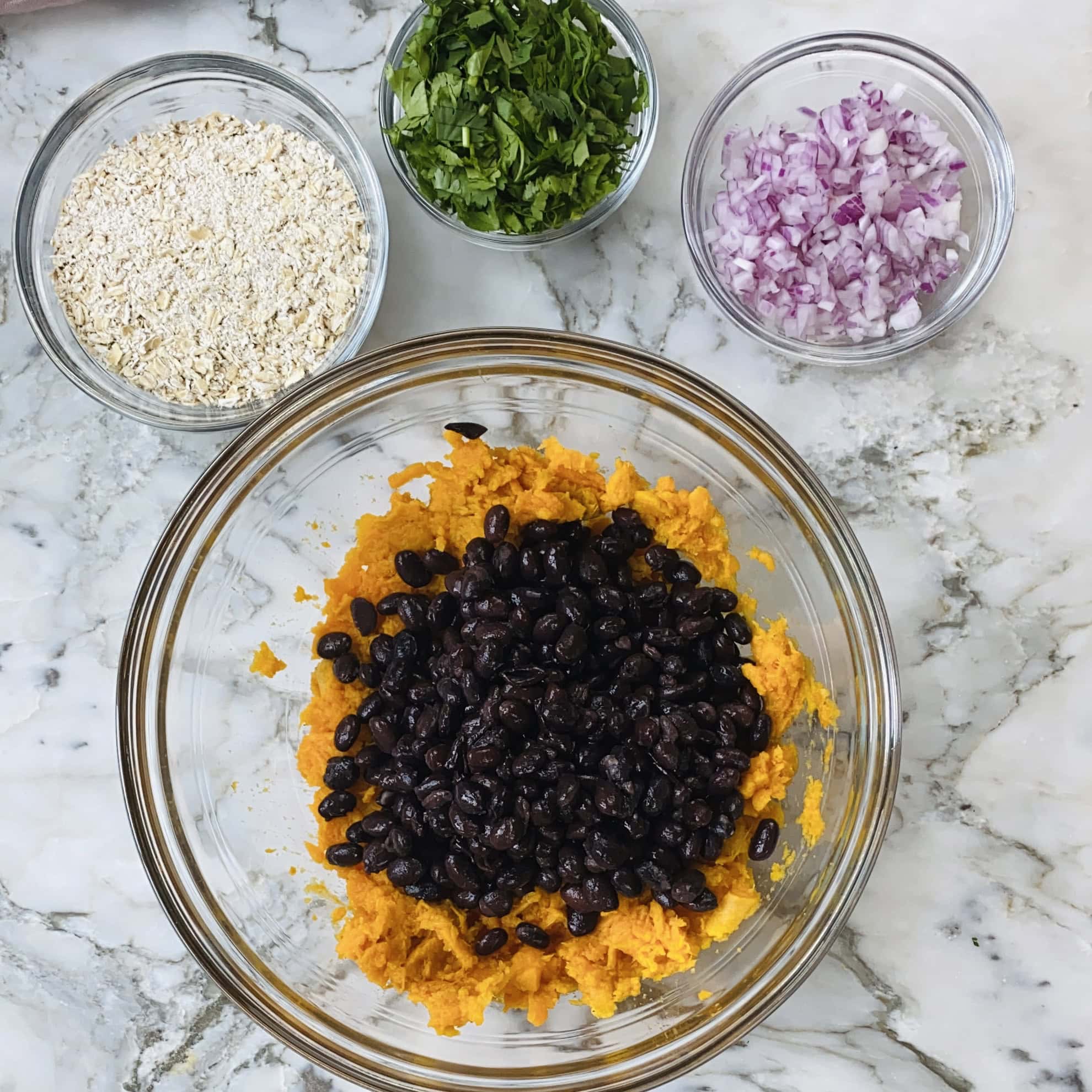 A glass bowl with mashed sweet potatoes and black beans, surrounded by bowls of oats, chopped parsley, and diced red onions on a marble surface.