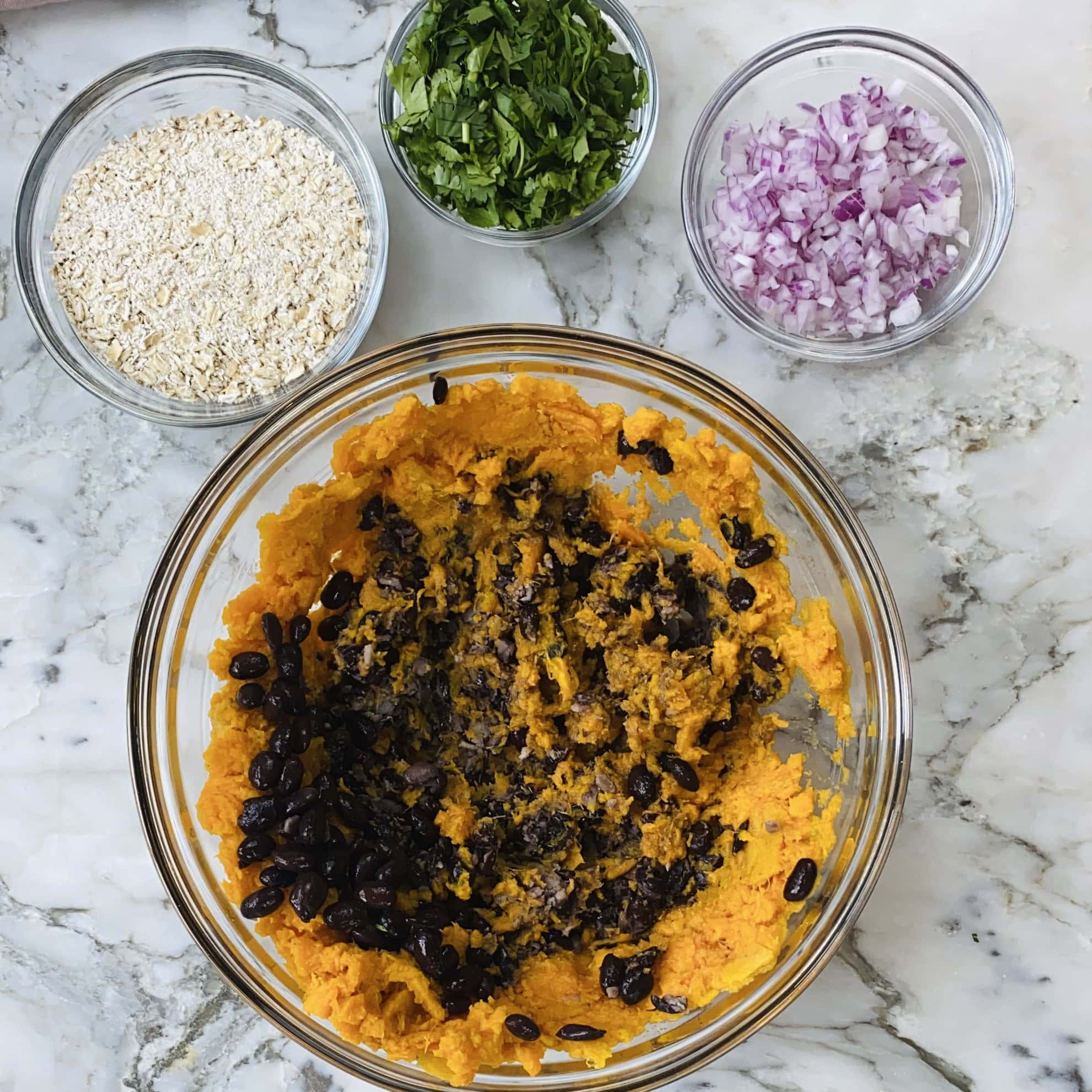 A bowl with mashed sweet potato and black beans, next to small bowls of chopped red onion, chopped herbs, and oats on a marble countertop.
