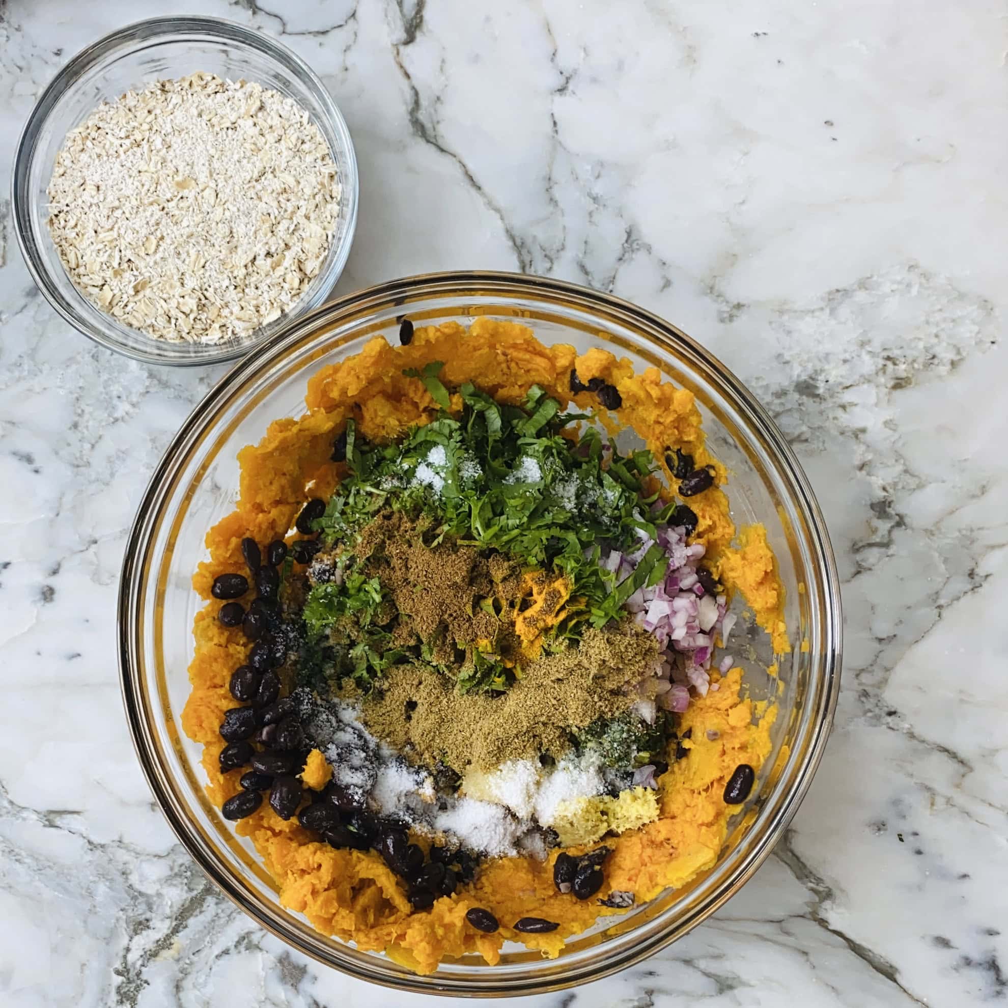 A glass bowl with mashed sweet potato, black beans, chopped red onion, herbs, and spices on a marble surface next to a smaller bowl of oats.
