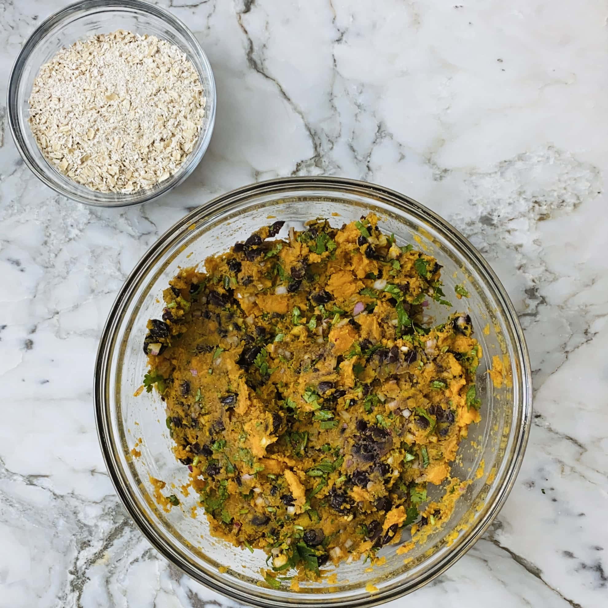 A glass bowl with a mixed vegetable mash containing herbs and beans sits on a marble surface next to a small bowl of oats.