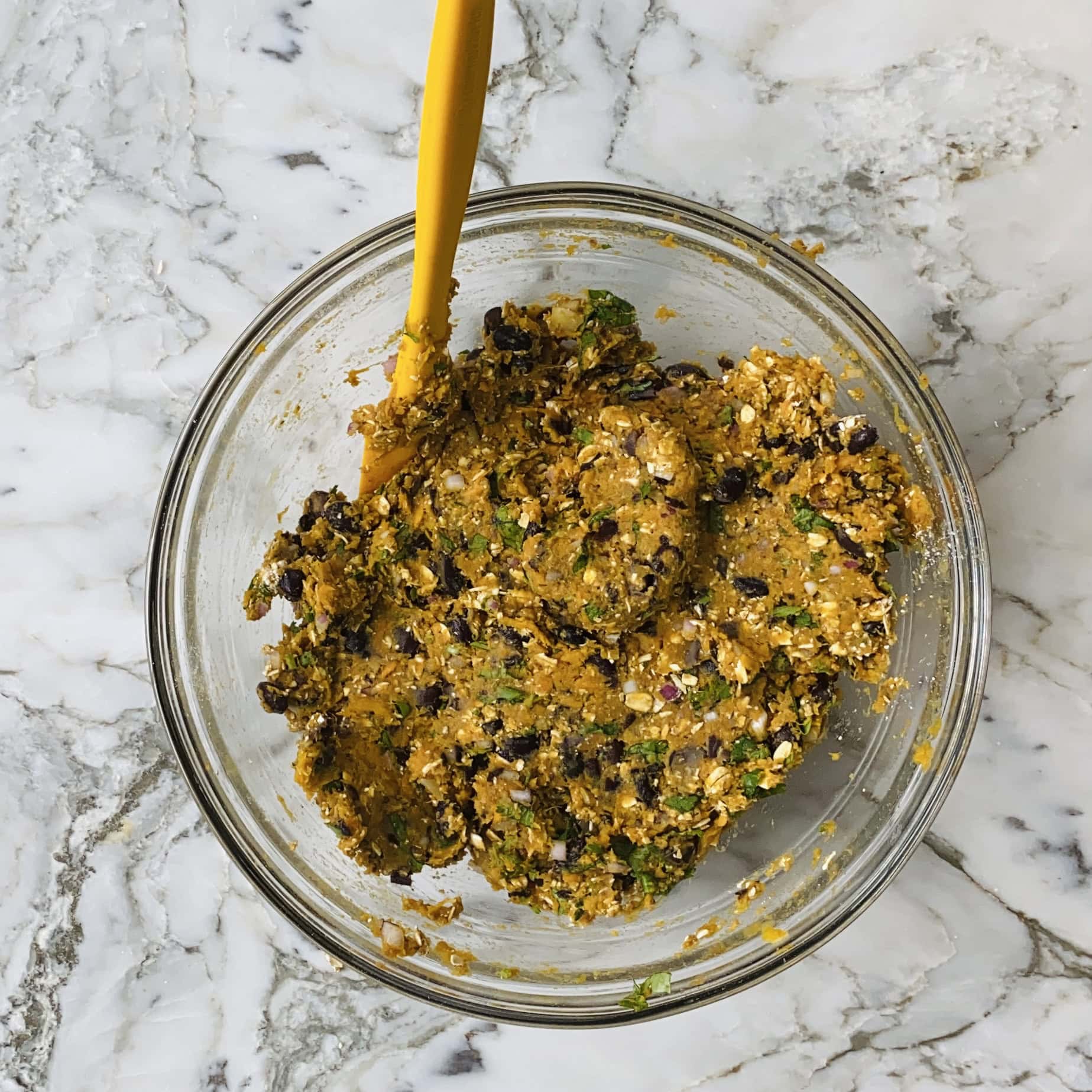 A glass bowl with a yellow spatula contains a mixture of black beans, oats, herbs, and other ingredients on a marble surface.