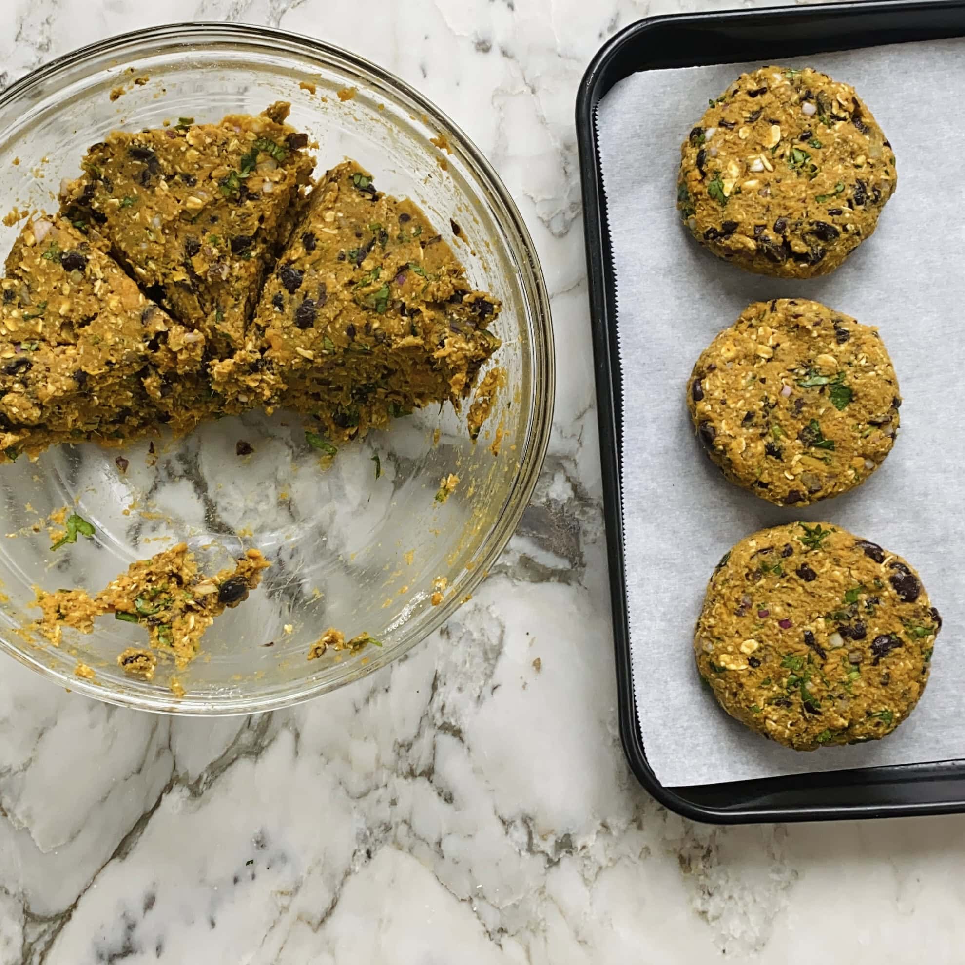 A glass bowl with mixed veggie burger patty dough sits on a marble counter next to a baking tray with three formed, uncooked patties on parchment paper.