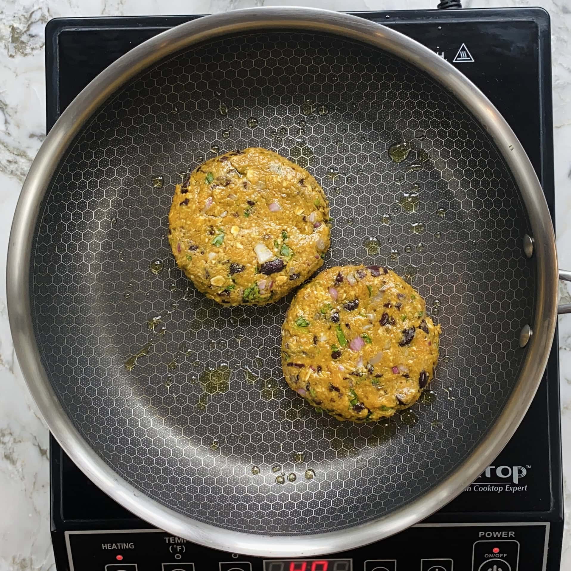 Two vegetable patties are cooking in a hexagonal-patterned frying pan on an induction cooktop.