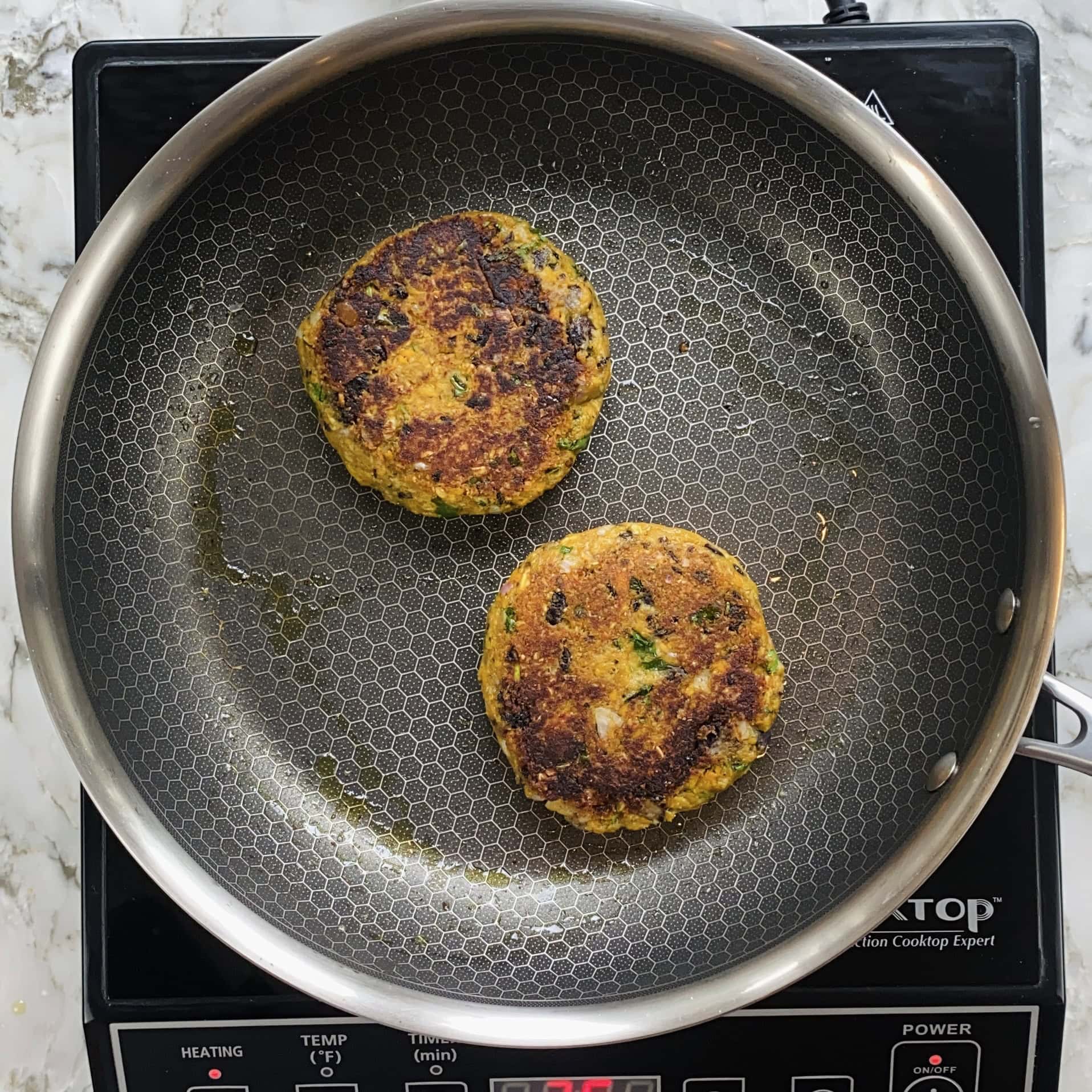 Two golden-brown vegetable patties cooking in a stainless steel pan on an induction cooktop.