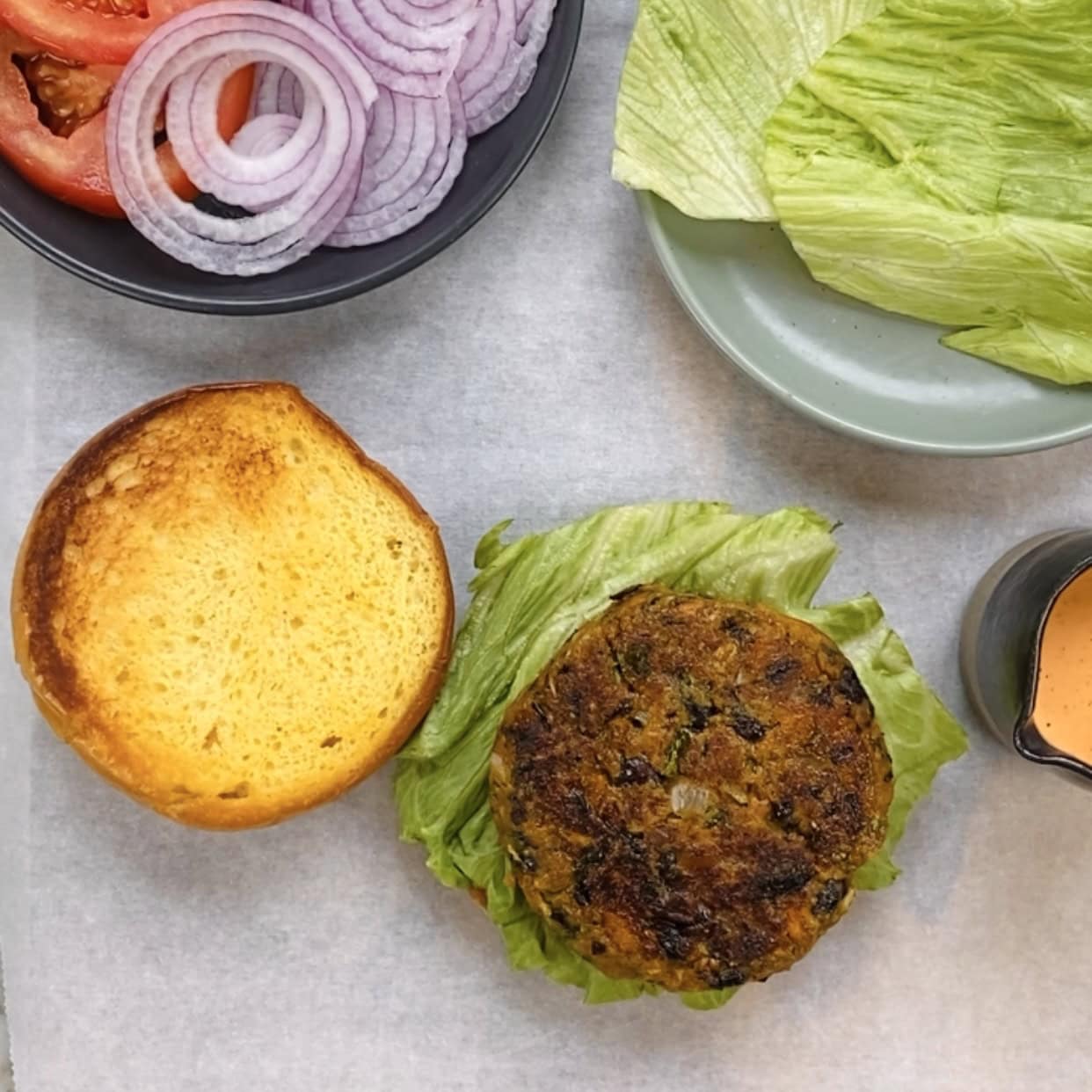 A veggie burger patty on lettuce with a toasted bun, sliced tomatoes, red onions, more lettuce, and a small bowl of sauce arranged nearby.