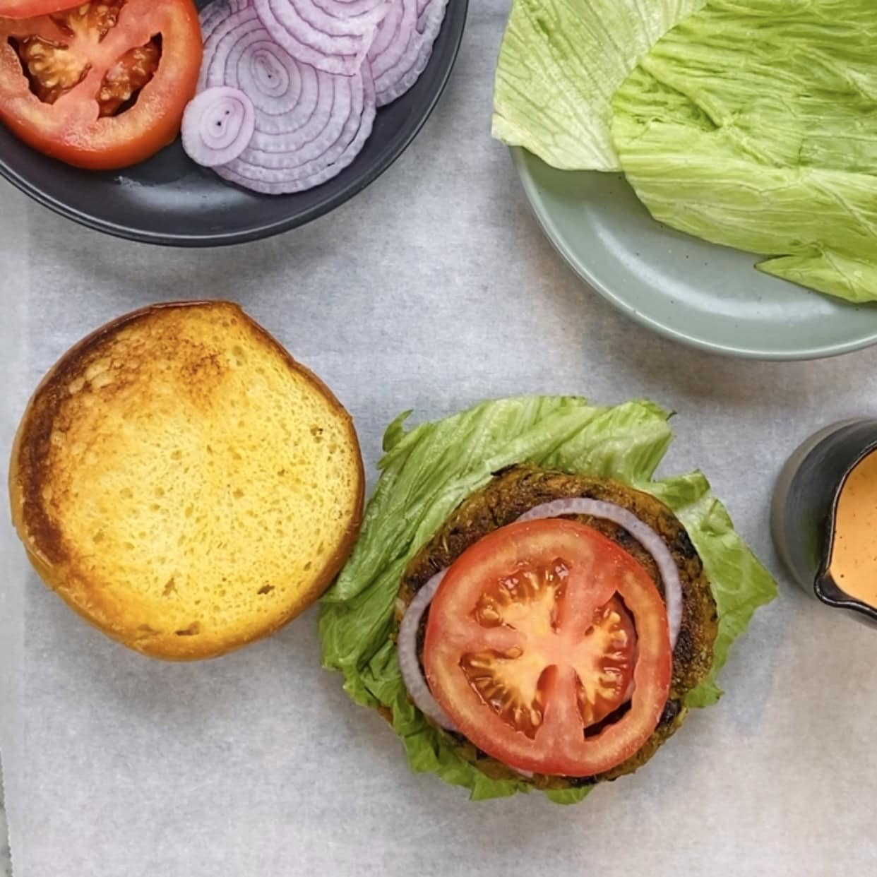 Ingredients for a burger, including a toasted bun, sliced tomato, red onion, lettuce, sauce, and a patty, arranged on a surface.