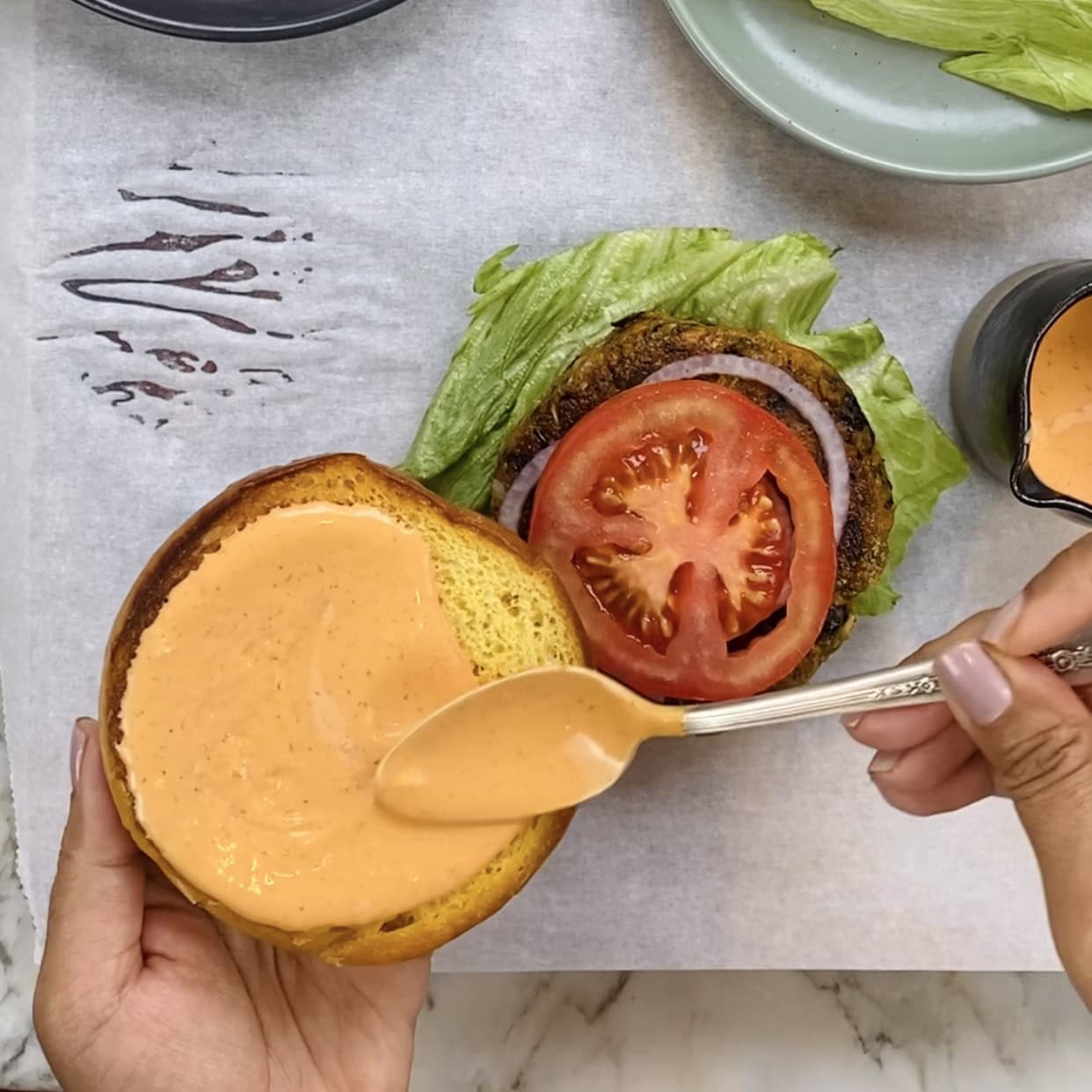 A hand spreads orange sauce on a burger bun next to a burger patty topped with lettuce, onion, and tomato.