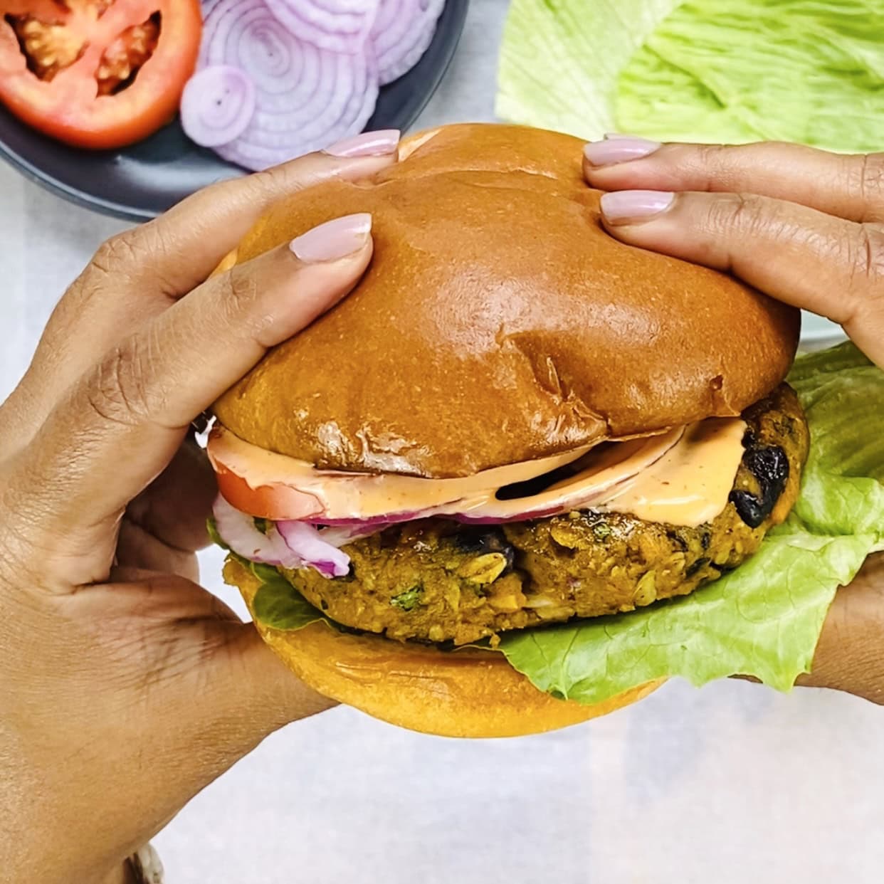 Hands holding a veggie burger with lettuce, tomato, red onion, and sauce in a brioche bun, with sliced vegetables in the background.