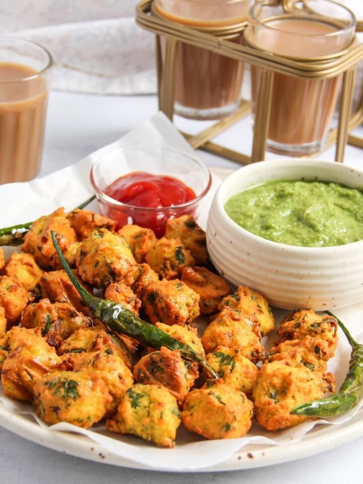 A plate of vegetable pakoras with green chili peppers, served with green chutney and ketchup, alongside glasses of chai tea.
