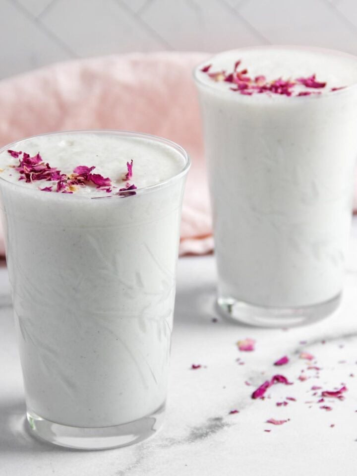 Two glasses of sweet lassi topped with dried rose petals, placed on a marble surface with a pink cloth in the background.
