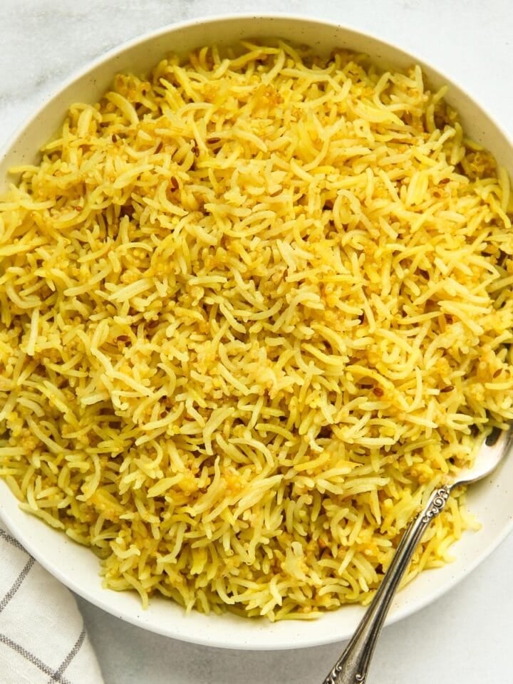 A white bowl filled with cooked yellow rice and quinoa, with a silver fork resting inside, placed on a white surface next to a white and gray checkered cloth.