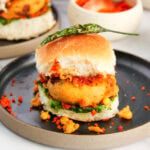 vada pav in a black plate with red chutney, chura and fried chili.