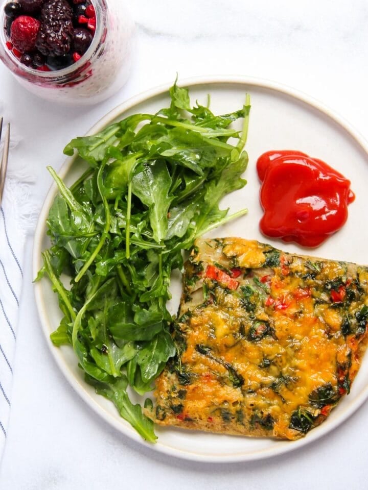 A plate with a slice of vegetable frittata, a serving of arugula, and two dollops of ketchup. A spoon, fork, and a glass of berry yogurt parfait are beside the plate.