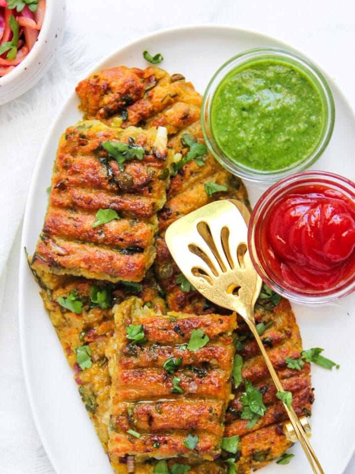 Grilled vegetable patties on an oval plate, garnished with herbs, served with green chutney and red sauce in small bowls, and a gold slotted serving spatula.