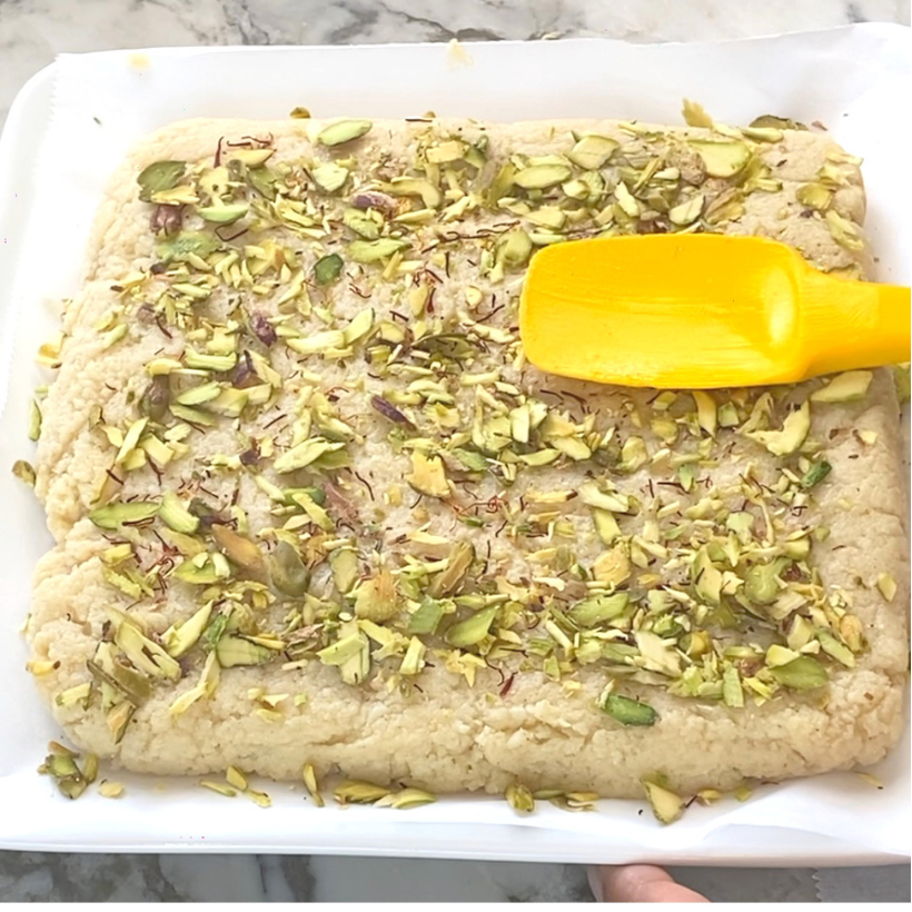 A square tray of pistachio-topped Indian sweet mixture being smoothed with a yellow spatula on a marble surface.