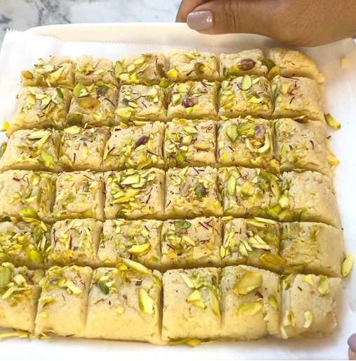 A hand holds a tray of square-shaped Indian sweets topped with chopped pistachios and saffron strands, arranged in neat rows.