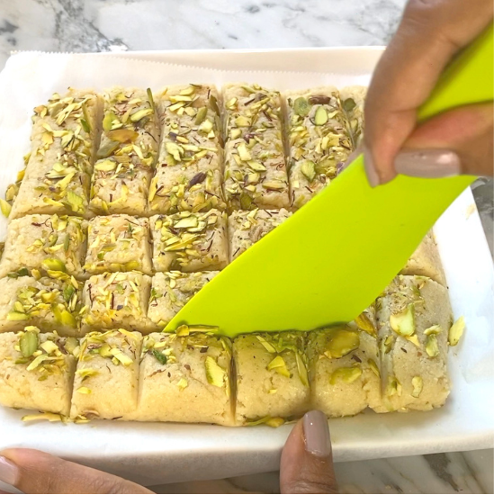 A lime green spatula cuts into a tray of square-cut pistachio-topped barfi sweets resting on parchment paper.